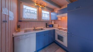 Ett litet, praktiskt kök med träpanel på väggarna, ett fönster med gardiner, en diskho, en spis och ett blått kylskåp.