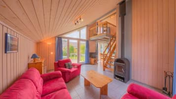 A cozy cabin interior with a wooden interior, a fireplace, a staircase to an upper deck, and a large window offering a view of the outdoors.