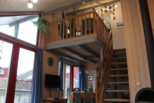 Un interior de madera con varios niveles, que incluye una escalera, un comedor con sillas y una mesa, y un ventanal grande.