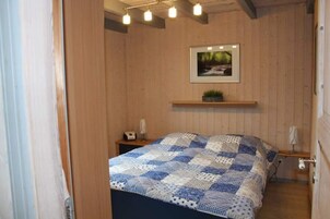 Un dormitorio con pared de madera, una cama con colcha azul y blanca, una mesita de noche pequeña, una mesita auxiliar, un cuadro enmarcado en la pared y un estante con una planta.