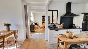 A modern kitchen with a black range hood, a dining area with a wooden table and chairs, and a view into a living room with a sofa and a coffee table.
