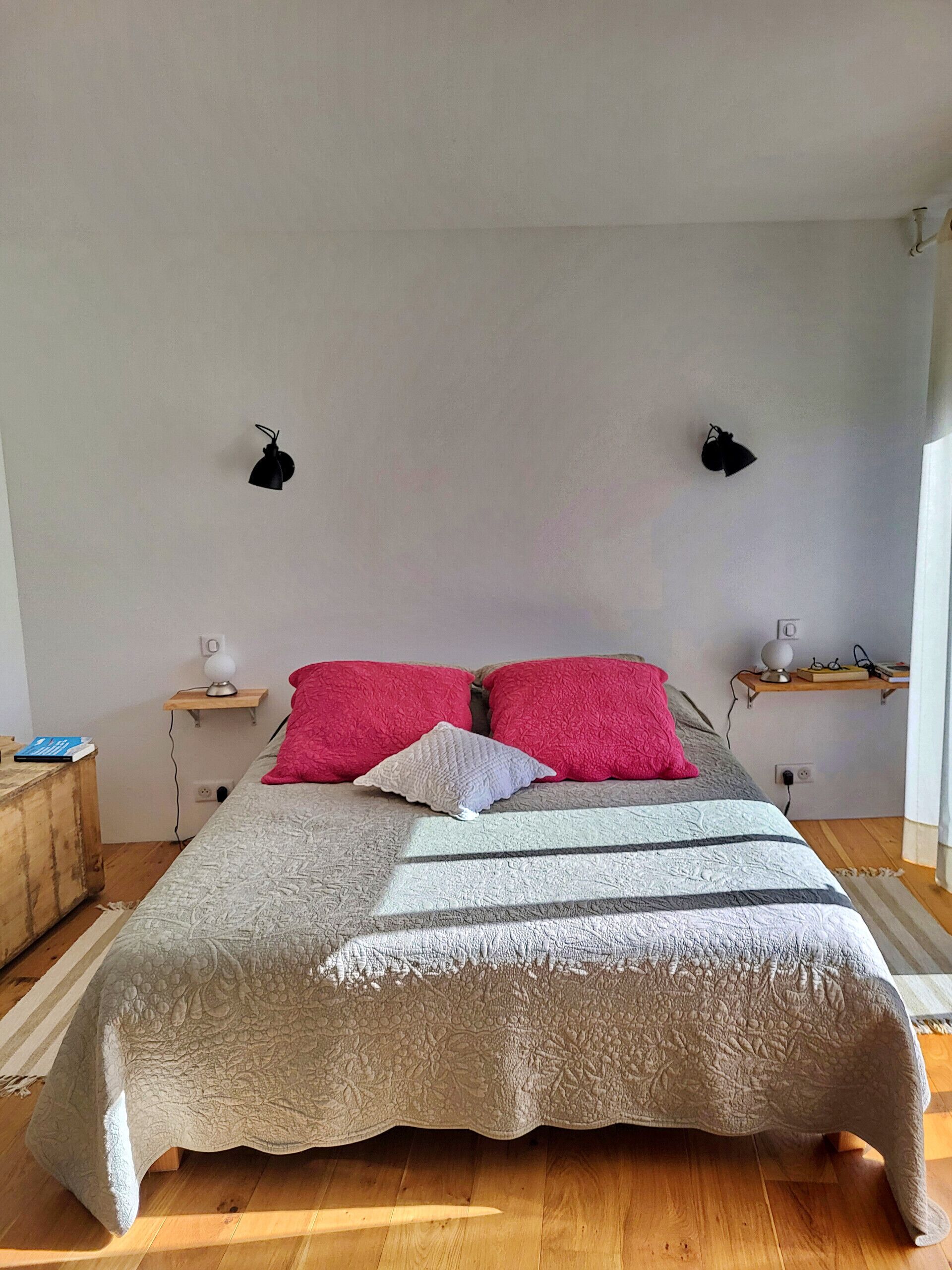 Un dormitorio con una cama grande, dos lámparas de noche, una mesita de noche, un estante con libros y gafas, y una ventana con cortinas.