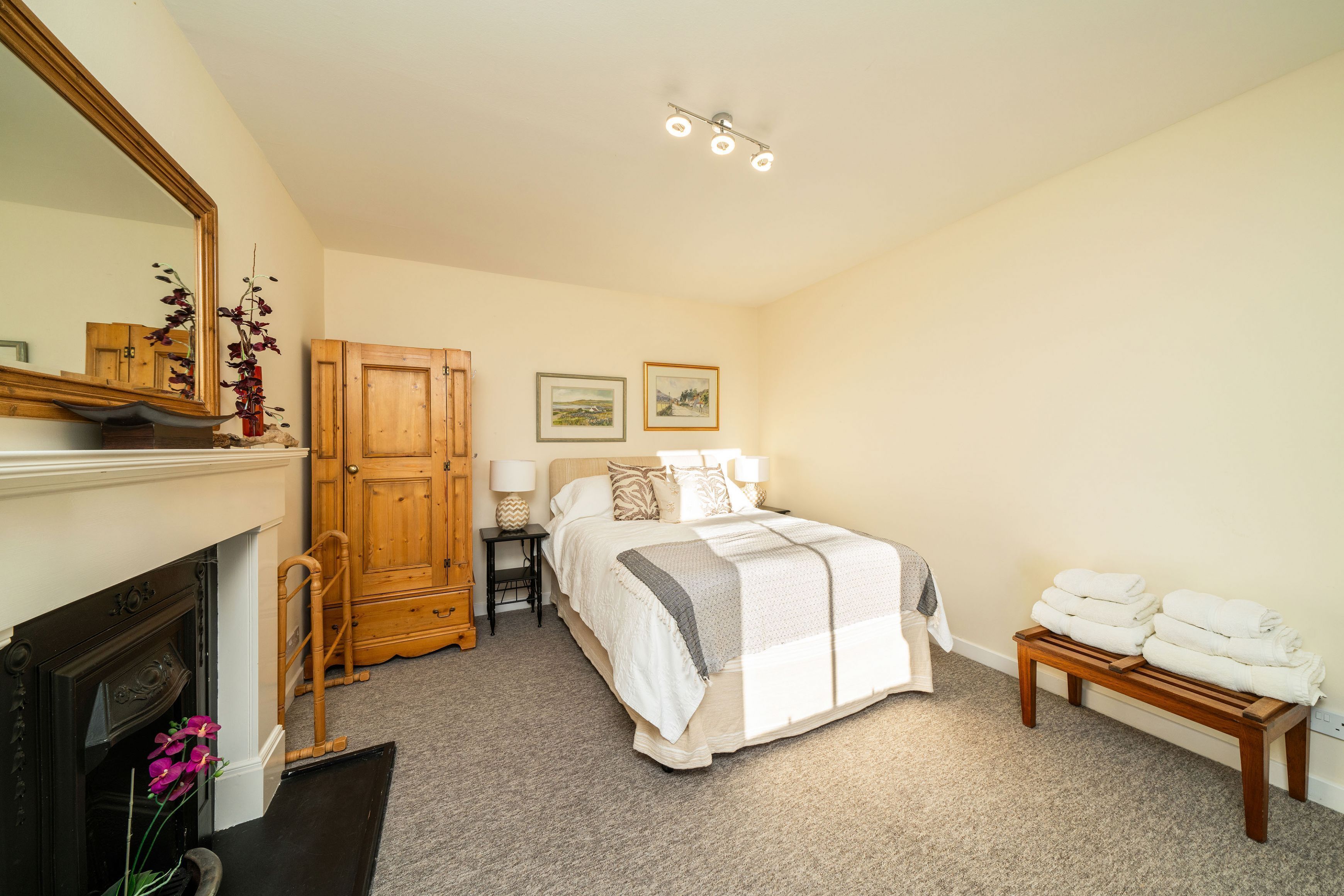 A bedroom with a bed, a wooden wardrobe, a nightstand, a lamp, a mirror, a fireplace, and a bench with towels.