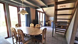 A dining area with a round wooden table and chairs, a ceiling fan, and a view of the outdoors through large windows. - Ferienhaus im Ferienpark Mirow (Mirow)