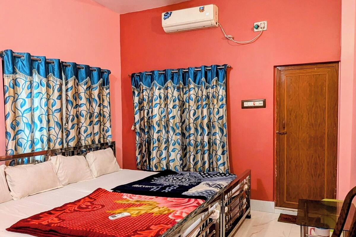A bedroom with a bed, a ceiling fan, an air conditioner, a wooden door, and a chair.