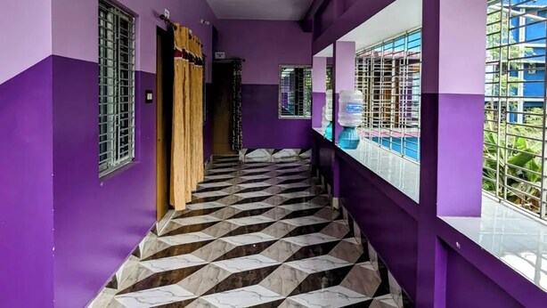 Un couloir aux murs violets et au sol en marbre à carreaux, qui mène à un balcon d’où l’on peut admirer un bâtiment bleu.