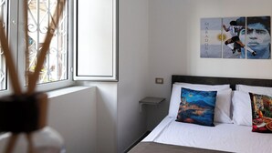 Een slaapkamer met een bed, een nachtkastje, een raam met metalen tralies en een muur met kunstwerken.