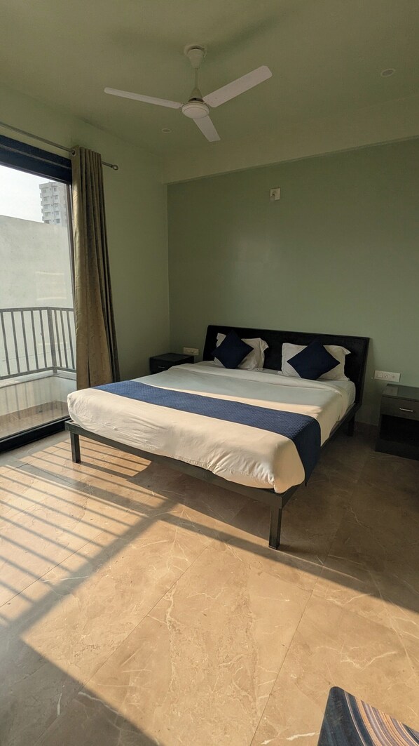 A hotel room with a bed, a ceiling fan, a nightstand, and a view of a building through a window. - MAHA RAAS A Unit of RAAS LEELA Hotel (Udaipur)