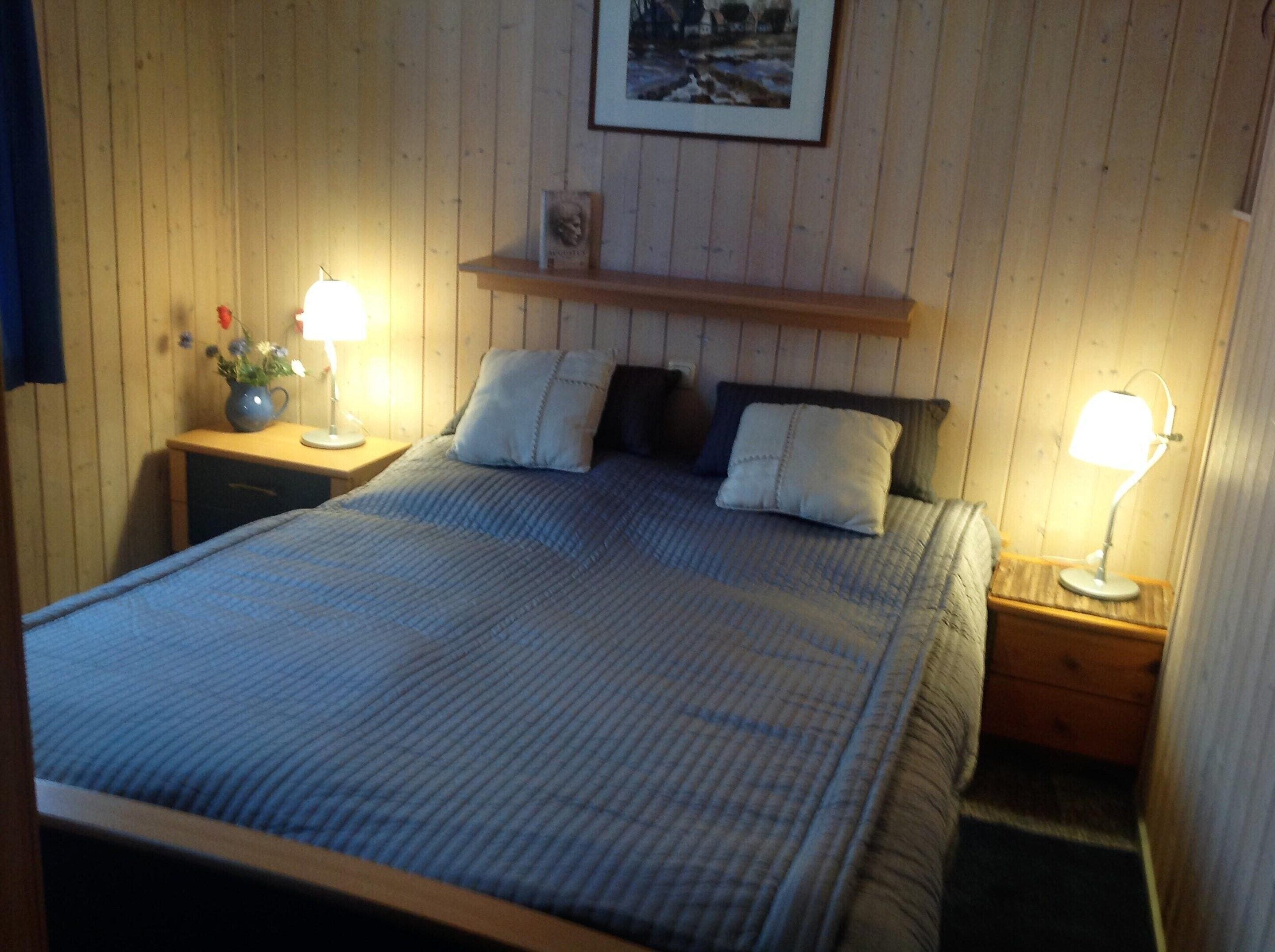 A neatly made bed with a blue quilt, two bedside lamps, a small shelf with a framed picture, and a nightstand with a vase of flowers.