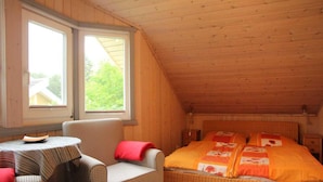 A room with a sloped ceiling, a bed with orange bedding, a checkered armchair, and a small table with a striped tablecloth. - Ferienhaus im Skandinavischen Stil (Mirow)