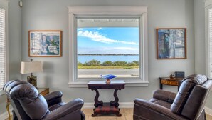 A room with a large window offering a view of a body of water and distant land. There are two leather chairs, a small table with a lamp, and two framed artworks on the walls. - Waterfront! Incredible Views, Watch the Dolphins Play, Perfect for Groups, $300 in Free Tickets! (St. Augustine)