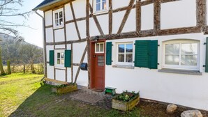 Una casa tradizionale a graticcio con una porta rossa, persiane verdi e un piccolo giardino con piante e una panchina di legno.