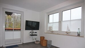 A room with a television, a window with blinds, a small shelf with decorative items, and a wicker basket. - Apartment in Wenkendorf for 6 Personen (Fehmarn)