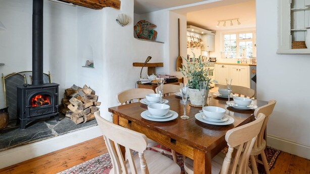 A cozy dining area with a stone fireplace, wooden dining table, and chairs. A kitchen is visible in the background.