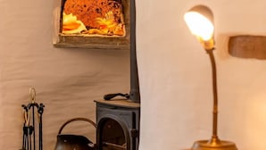 A rustic room with a wood-burning stove, a basket of firewood, and a table with a lamp.