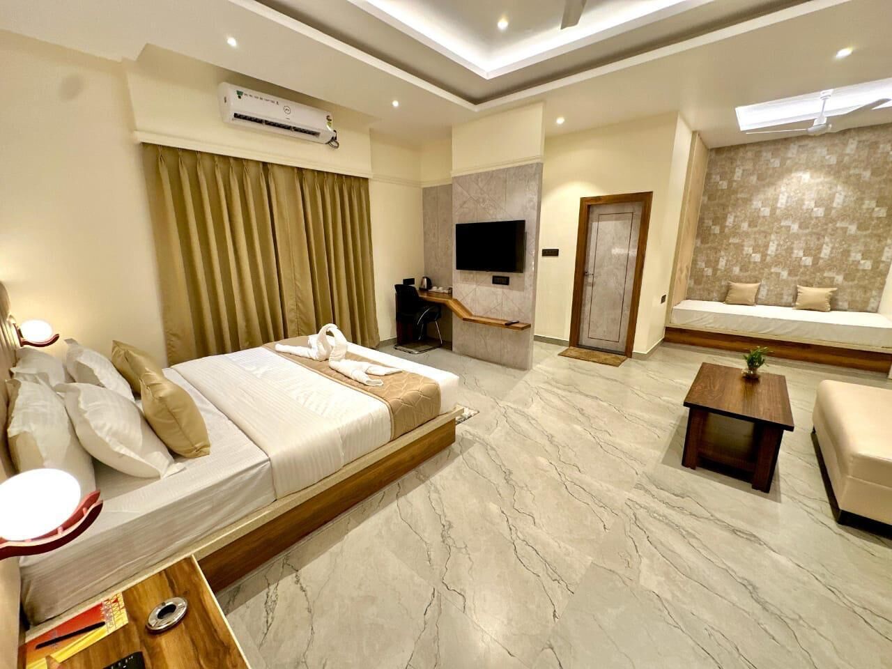 A modern hotel room with a large bed, a TV mounted on the wall, a seating area with a sofa and a small table, and a marble floor.