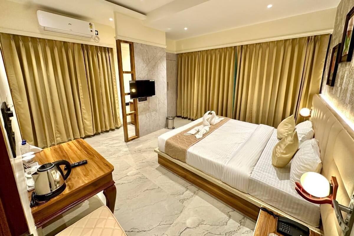 A hotel room with a bed, a wooden side table with a kettle, a TV, and a minibar.