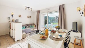 A dining area with a table set for two, a sofa, a TV, and a view of a pool and trees.