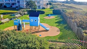 A playground with slides and climbing structures, surrounded by a fence and a building with balconies in the background.