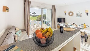 A modern living room with a large window, a dining area with a fruit bowl, and a TV on a stand.