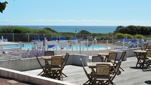 A poolside area with lounge chairs and tables, a pool, and a view of the ocean.
