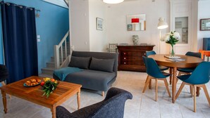 Ein modernes Wohnzimmer mit einer Couch, einem Holzesstisch und Stühlen. Eine Treppe führt zu einer weiteren Ebene, eine blaue Wand und ein rotes Pendelleuchten sind zu sehen.