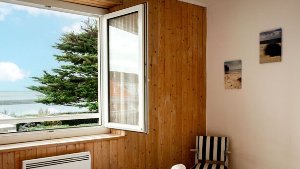 Ein Schlafzimmer mit Holzwand, einem Bett mit weißen Leinentüchern, einem Fenster mit Blick auf Bäume und Wasser sowie einem Sessel mit gestreiften Kissen.