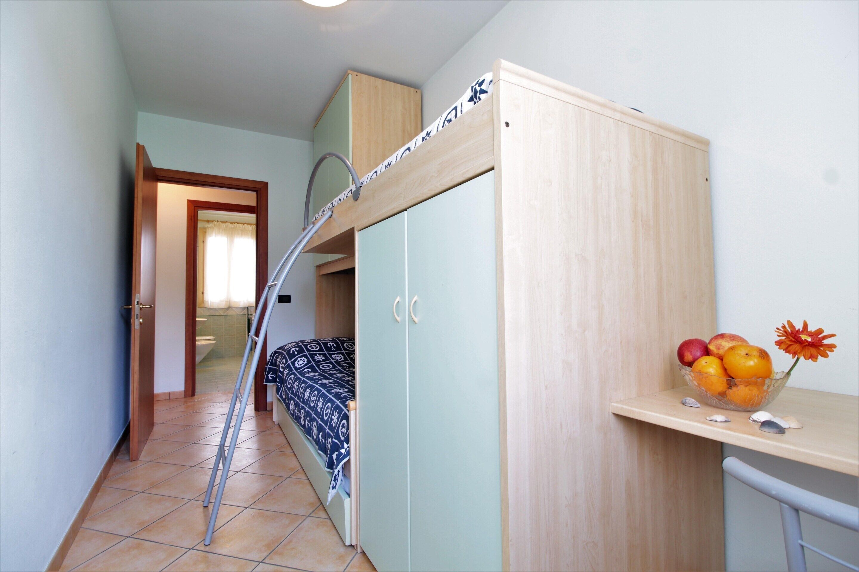 A compact room with a bunk bed, a wooden wardrobe, a small table with fruit, and a chair.