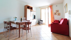 A living room with a red sofa, a dining table with chairs, a kitchen area, and a sliding glass door leading to a balcony. - Belvilla by OYO Appartamento Rosso (Jesolo)