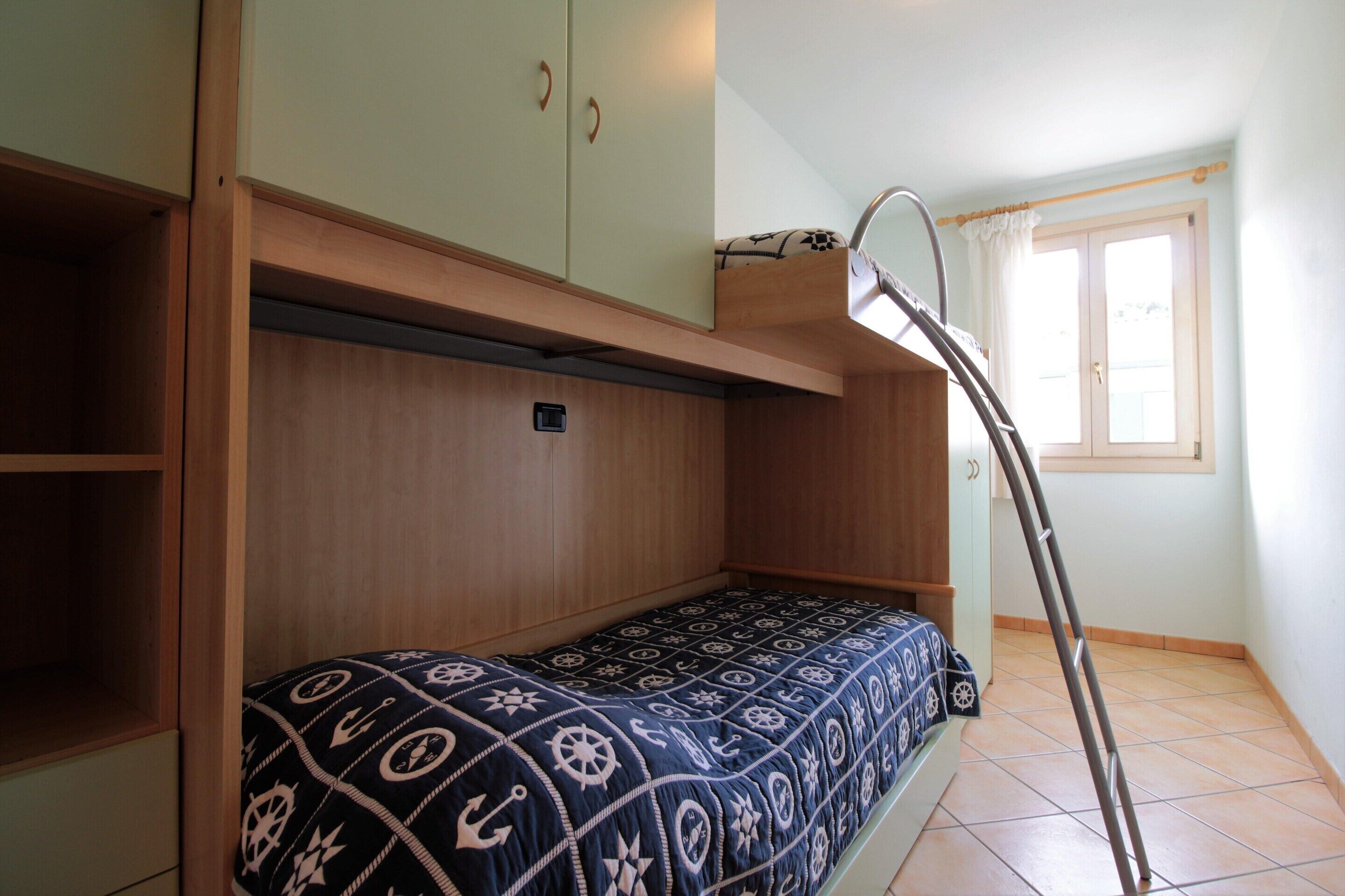 A bunk bed with a patterned bedspread, a wooden shelf, and a window with curtains.
