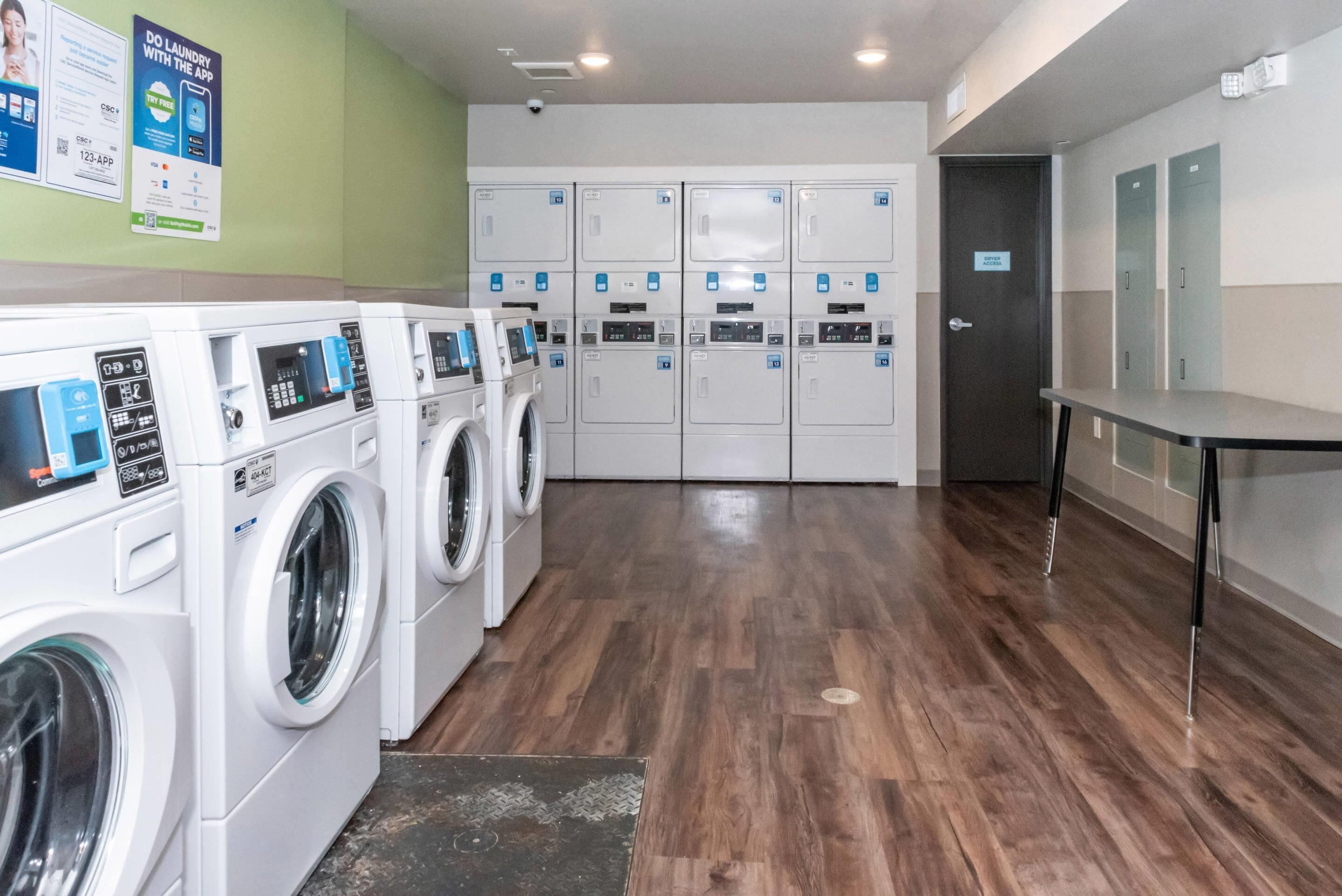 Une buanderie équipée de plusieurs machines à laver et de plusieurs sèche-linge, avec un panneau fixé au mur et une porte portant l’inscription « ACCÈS AUX SÈCHE-LINGE ».