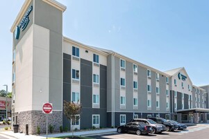 Un hotel de varios pisos con un cartel llamativo de 'Woodspring Suites', un estacionamiento con varios autos y un cartel de 'NO PASAR'.