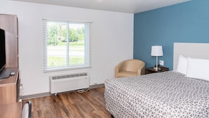 A hotel room with a bed, a chair, a desk with a TV and remote, a window with blinds, and a wall heater.