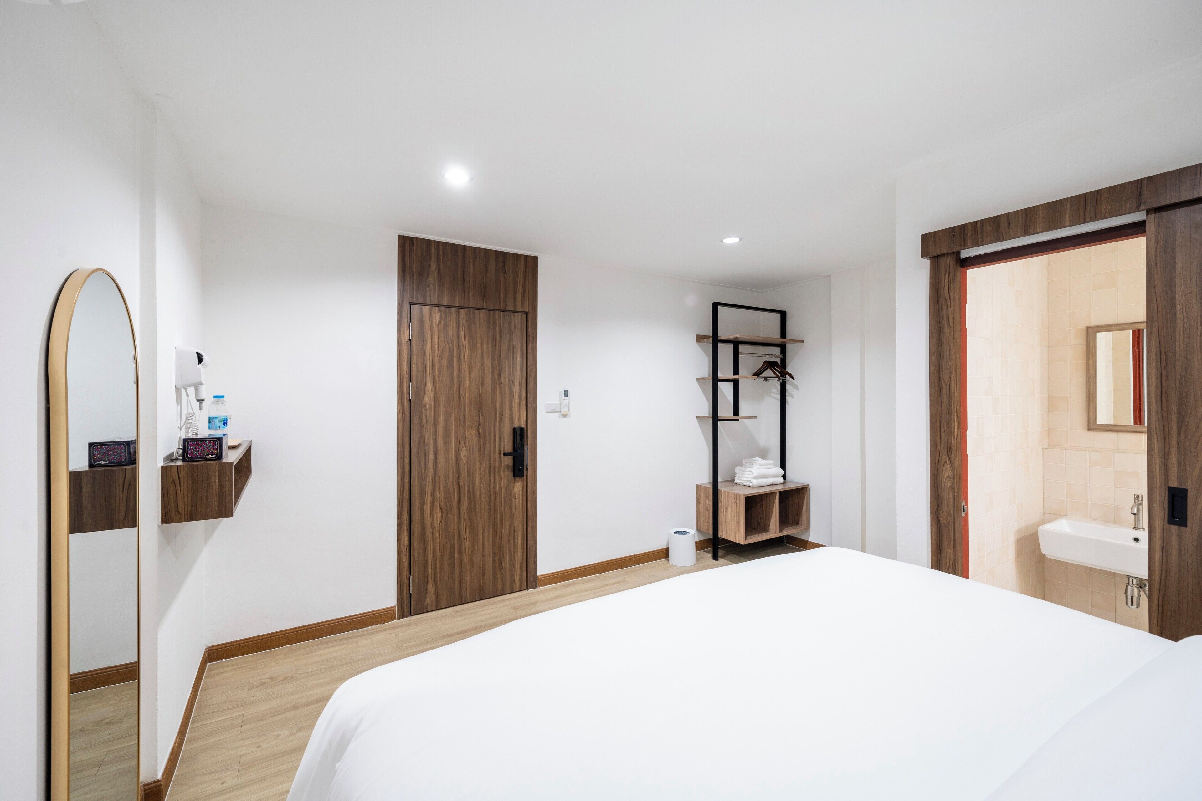 A modern hotel room with a large bed, a wooden wardrobe, a small bathroom with a sink, and a shelf with toiletries.