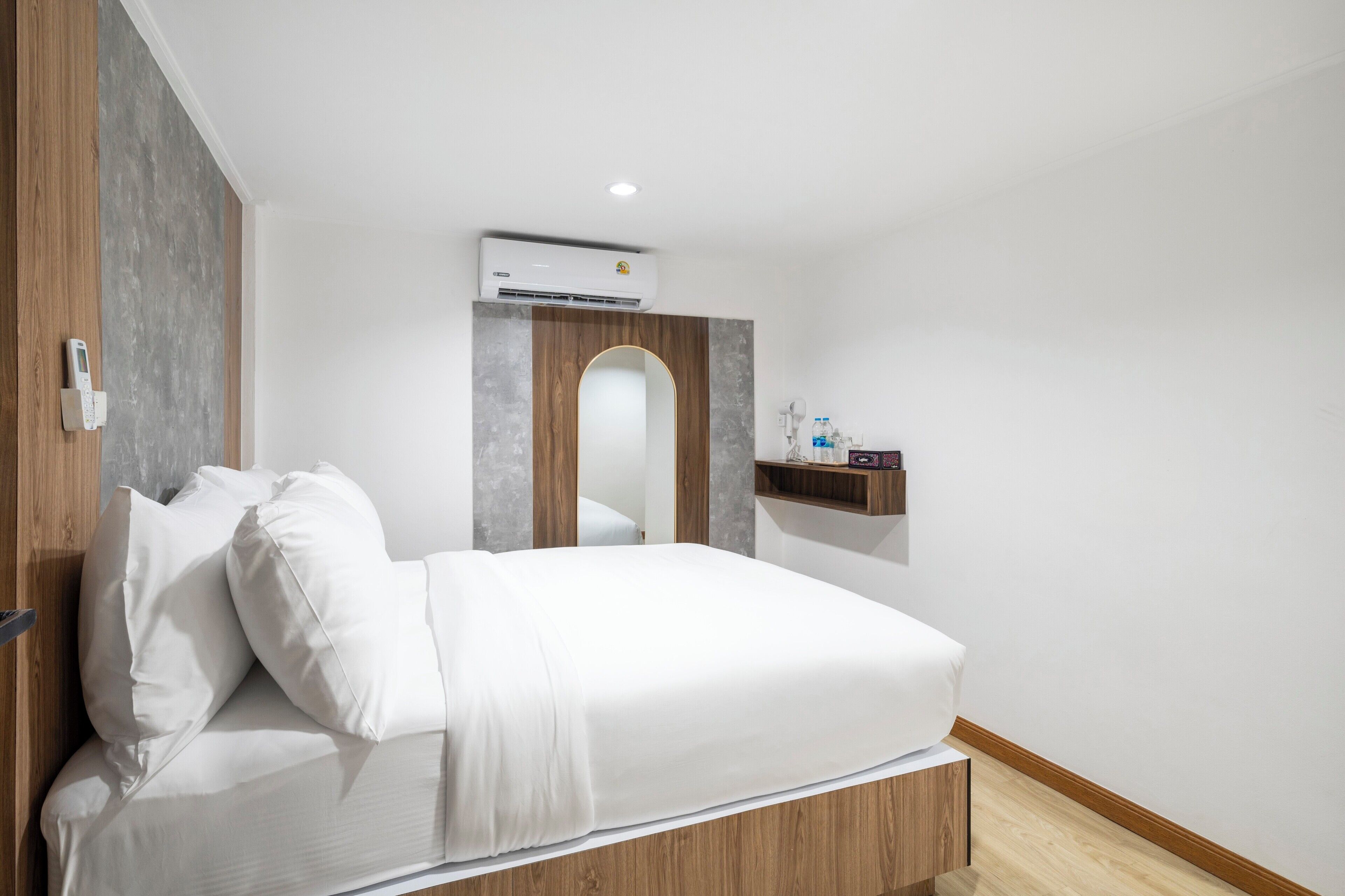 A modern hotel room with a wooden bed frame, white bedding, a mounted air conditioner, a small shelf with toiletries, and a wooden door.