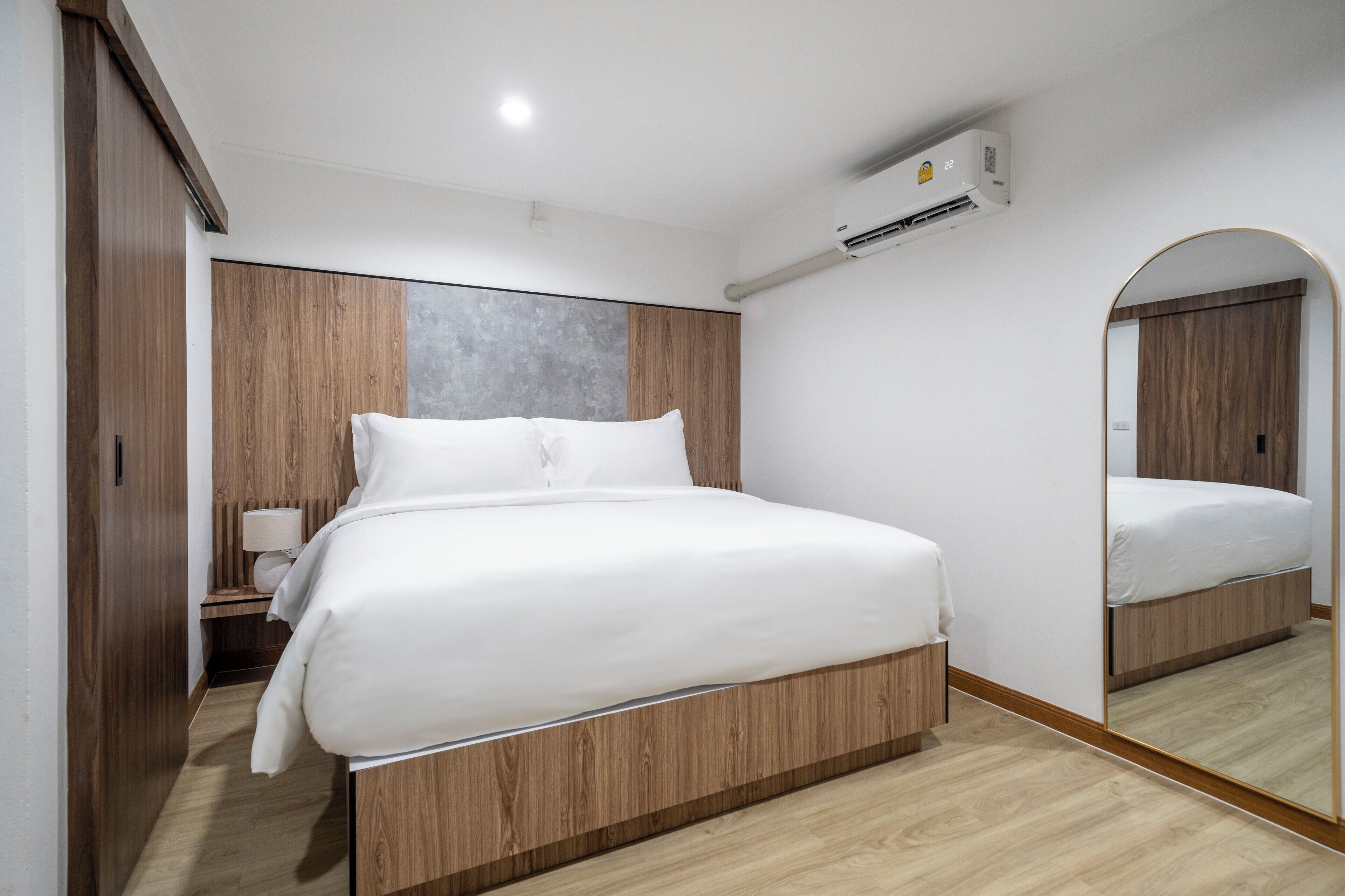 Un dormitorio moderno con una cama grande, cabecera de madera, una mesita de noche con lámpara, un espejo y un equipo de aire acondicionado.