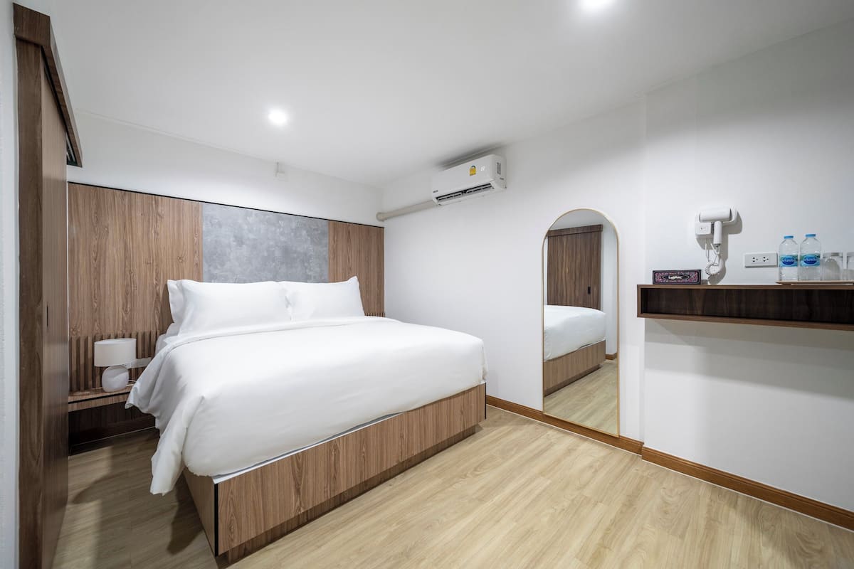 A modern hotel room with a large bed, built-in wooden headboard, a bedside table with a lamp, an air conditioning unit, a mirror, and a shelf with bottled water.