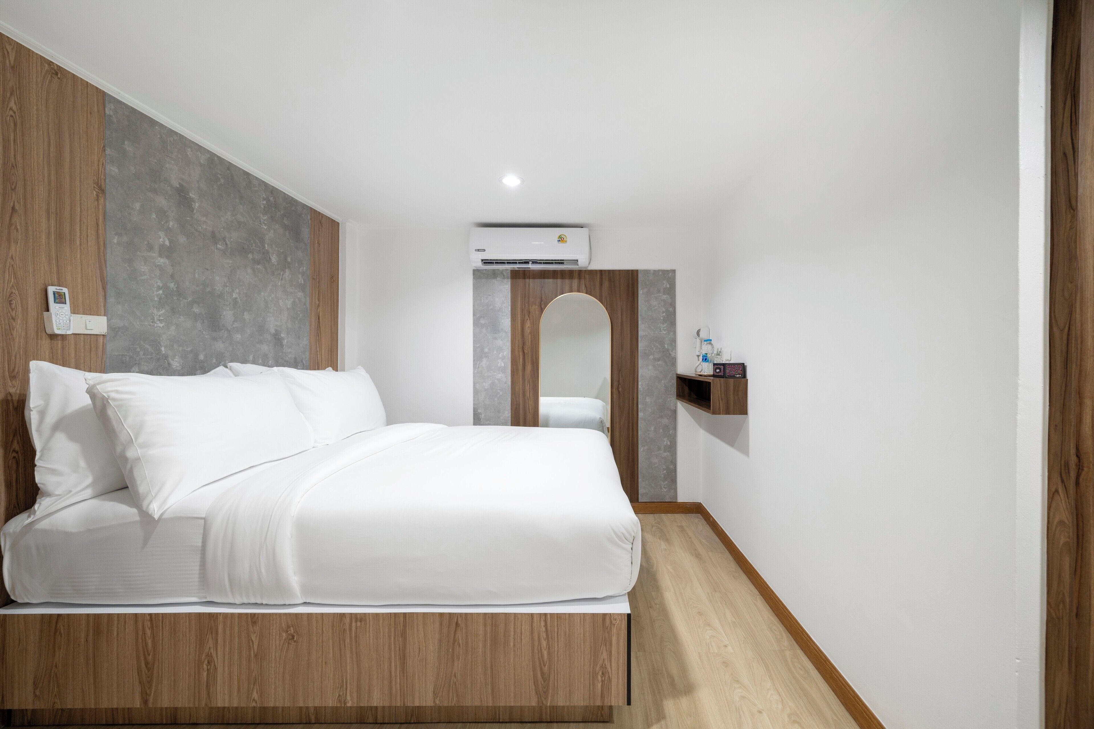 A modern bedroom with a wooden headboard, a large bed with white bedding, a bedside table, a wall-mounted air conditioner, and a small shelf with toiletries.