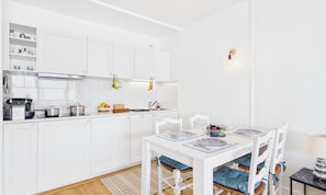 Une cuisine moderne avec des meubles blancs, une table à manger avec des sets de table bleus et une petite table d’appoint avec une lampe.