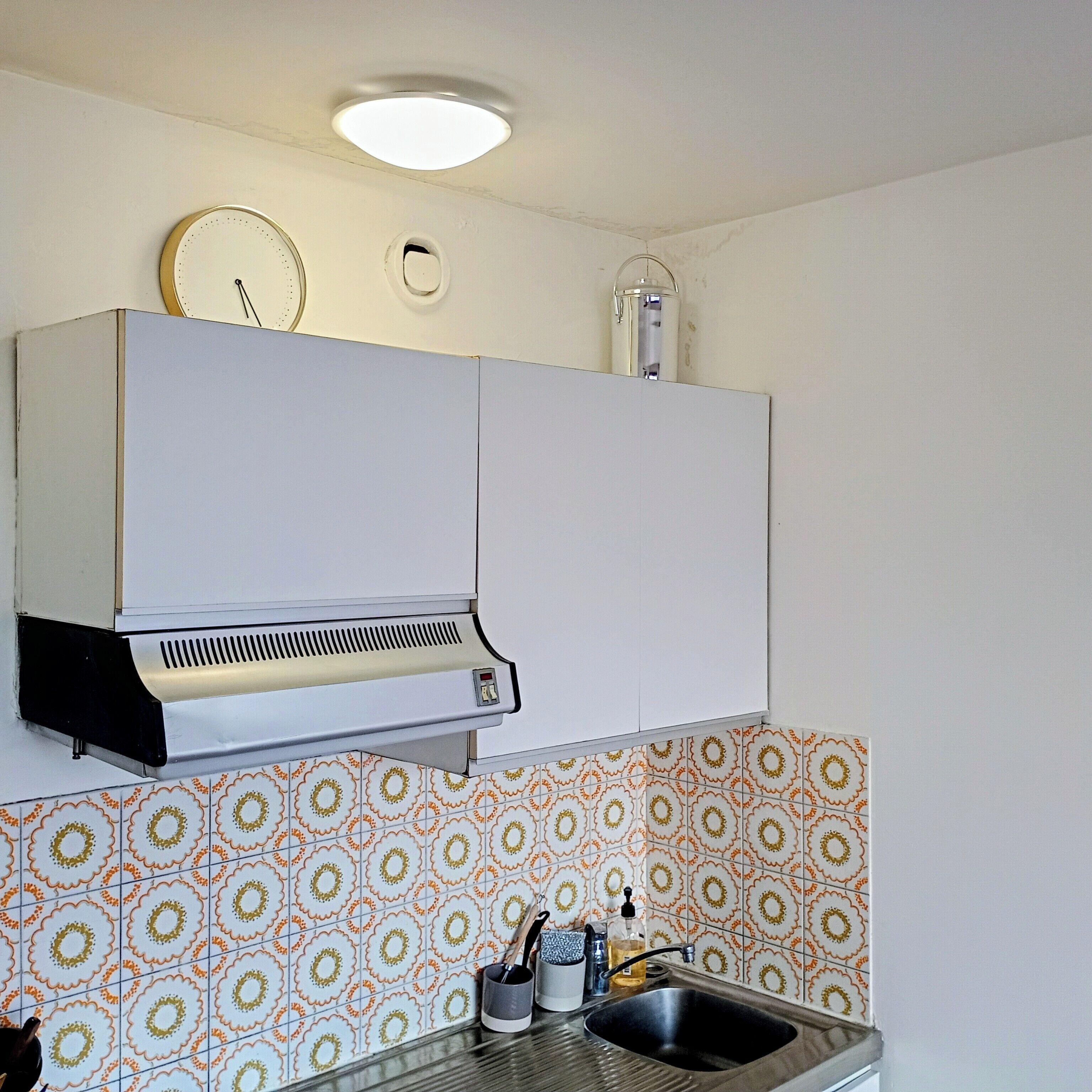 Een keuken met witte kastjes, een inbouwafzuigkap, een spoelbak en betegelde achterwand met een patroon.