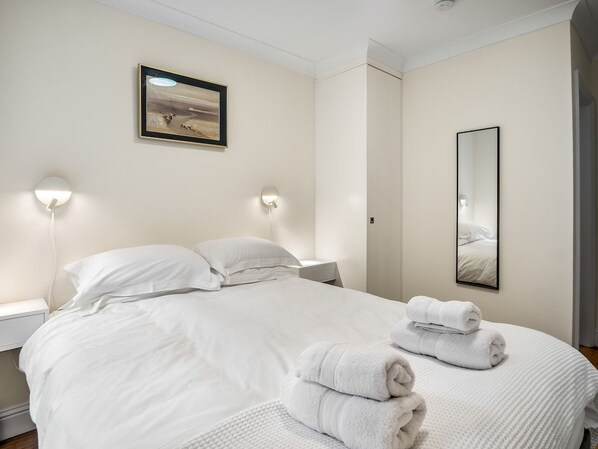 A neatly made bed with white linens and towels, a bedside lamp, a framed picture on the wall, and a mirror.