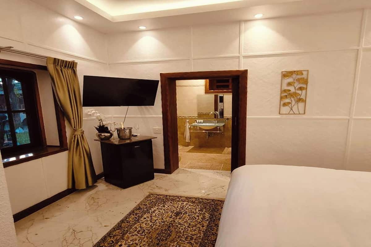 Ein Hotelzimmer mit einem Bett, einem Fernseher, einem kleinen Tisch mit einer Vase, einem Fenster mit Vorhängen, einem durch eine geöffnete Tür einsehbaren Badezimmer und einem gemusterten Teppich.