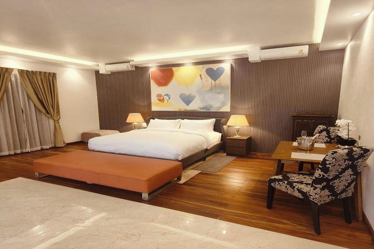 A hotel room with a bed, bedside tables, a chair, a small table, and a painting on the wall.