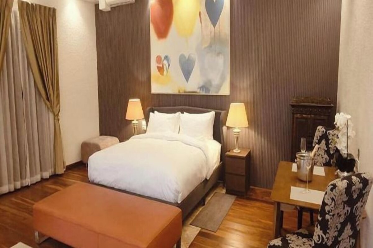 A hotel room with a bed, bedside tables, a small table with a chair, and a painting on the wall.
