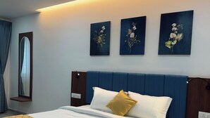 Una cama bien hecha con cabecera azul, ropa de cama blanca y colcha amarilla. Hay tres cuadros colgados en la pared sobre la cama, un ventilador de techo y un espejo en la pared.