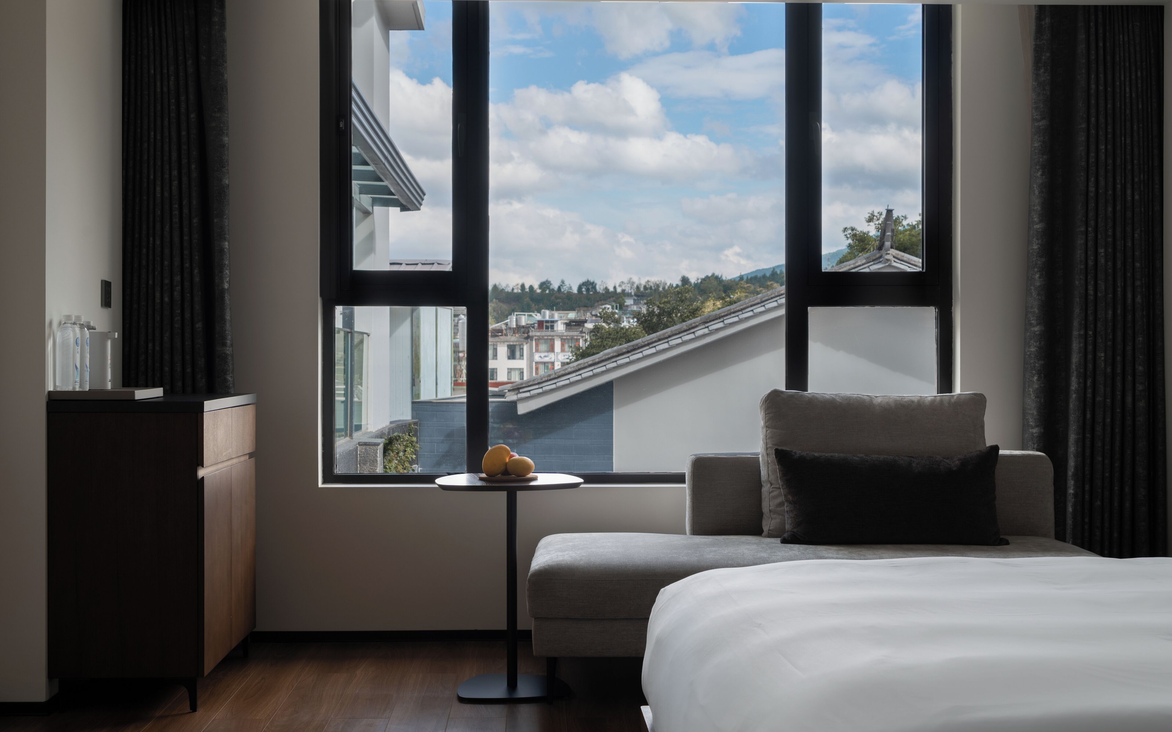 A hotel room with a bed, a small table with a fruit bowl, a chair, and a view of buildings through large windows.