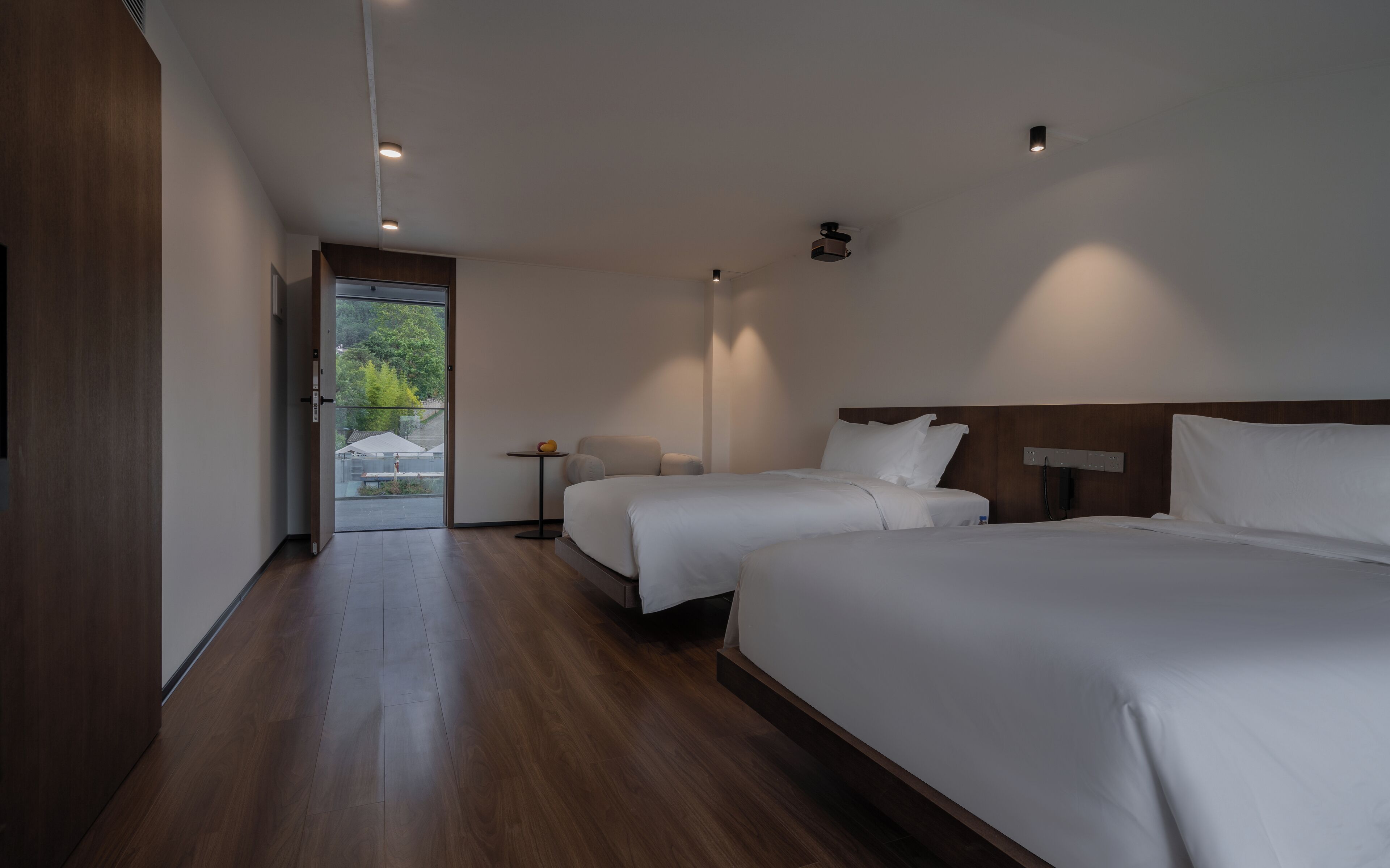 A modern hotel room with two single beds, a small bedside table, and a view of the outdoors.