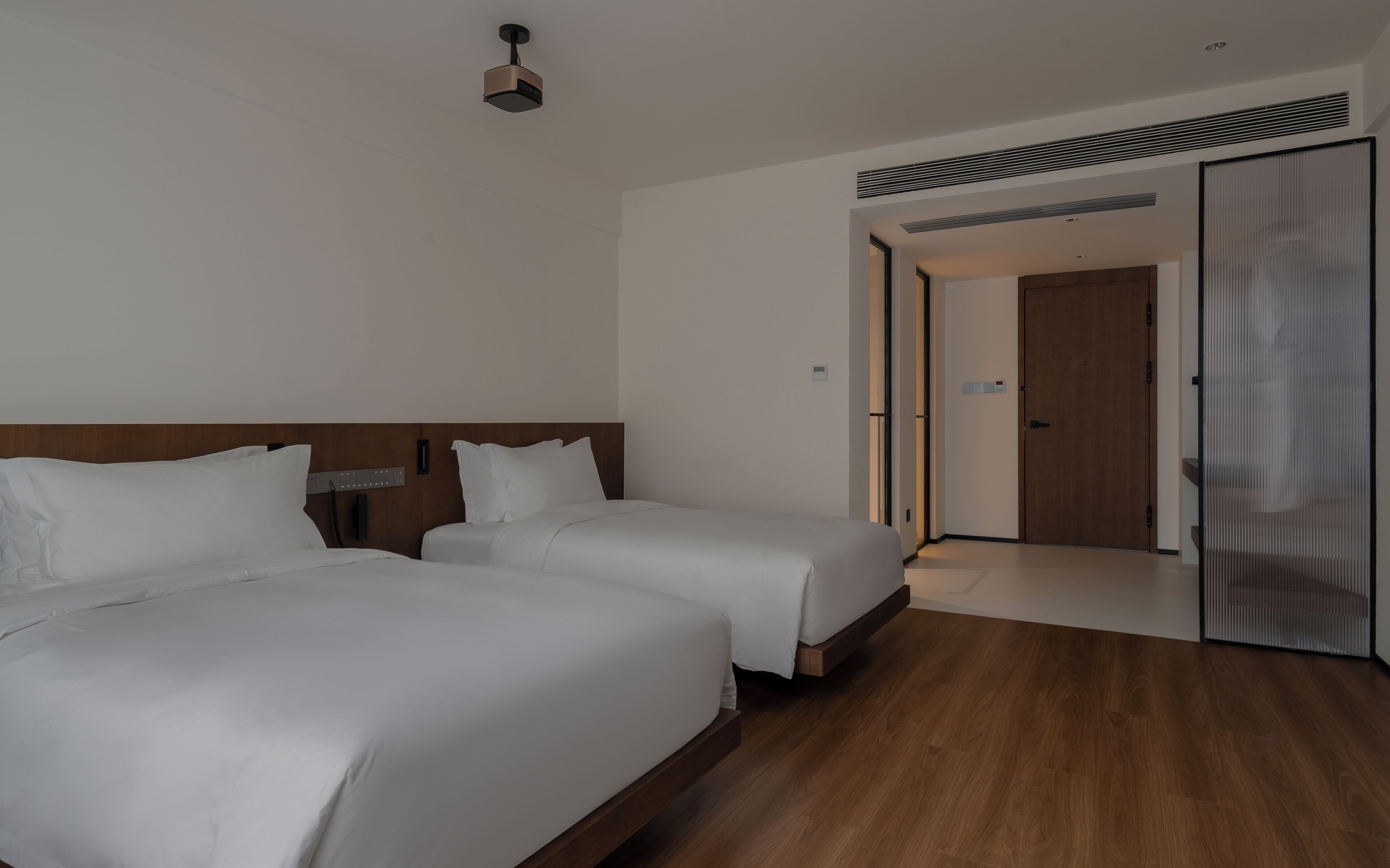 A hotel room with two beds, a wooden headboard, a ceiling projector, and a sliding door leading to another room.