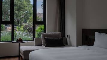 A hotel room with a large window, a bed, a chair, and a small table with fruit.