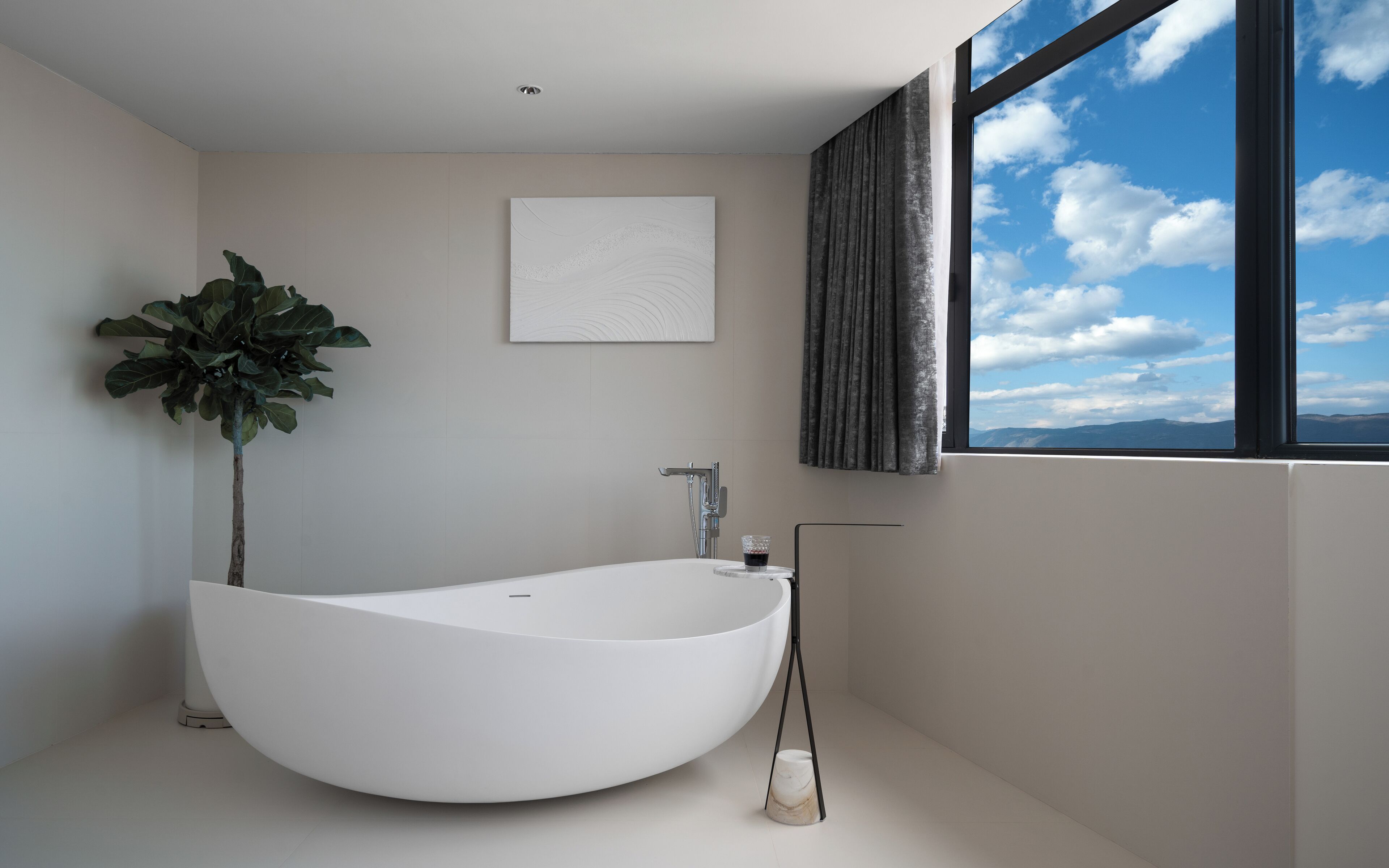 Un baño moderno con una bañera independiente, una planta en maceta, una obra de arte enmarcada y vistas del cielo a través de una ventana.